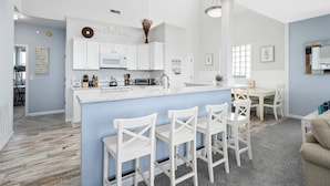 A modern kitchen with white cabinets, a central island with bar stools, and a dining area with a white table and chairs. - Sunset Overlook, Spacious bay side top-floor condo with amazing views  (Ocean City)