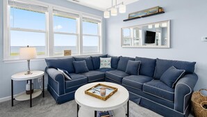 A living room with a blue sectional sofa, a round coffee table, a side table with a lamp, and a large window with blinds. - Sunset Overlook, Spacious bay side top-floor condo with amazing views  (Ocean City)