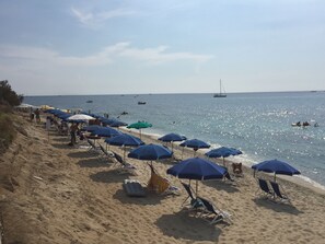 Een strand met rijen ligbedden en parasols, een zandstrand en boten op het water.