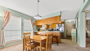 A dining area with a wooden table and chairs, a kitchen with wooden cabinets, and a hallway with a door. - Condo steps to beach w/ daily attraction perks (Panama City Beach)