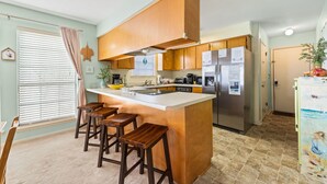 A kitchen with wooden cabinets, a central island with stools, a refrigerator, and a window with blinds. - Condo steps to beach w/ daily attraction perks (Panama City Beach)