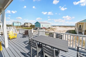 A wooden deck with a table and chairs, overlooking a beach with houses in the background. - 3BR/2BA Fort Morgan Home | Sleeps 8 | Gulf Views & Bunkroom Fun (Gulf Shores)