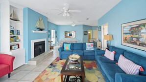 A living room with a blue sofa, a red armchair, a fireplace, and a ceiling fan. - La De Da | 3BR/2BA Fort Morgan Home | Sleeps 8 | Gulf Views & Bunkroom Fun (Gulf Shores)