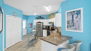 A kitchen with white cabinets, a granite countertop, and a dining area with wicker chairs. - La De Da | 3BR/2BA Fort Morgan Home | Sleeps 8 | Gulf Views & Bunkroom Fun (Gulf Shores)