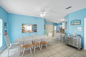 A dining area with a wooden table and white chairs, a ceiling fan, and a colorful painting on the wall. - 3BR/2BA Fort Morgan Home | Sleeps 8 | Gulf Views & Bunkroom Fun (Gulf Shores)