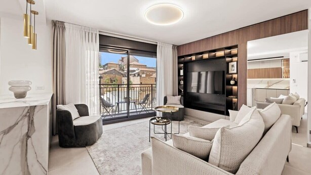A modern living room with a large TV, comfortable seating, a kitchen area, and a balcony with a city view. - Luxury 3 BDR  In The Heart Of Jerusalem By Isrentals (Jerusalem)