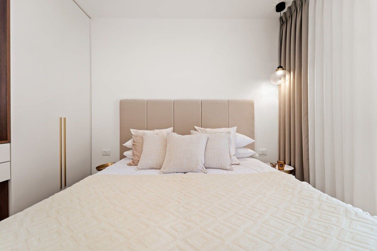 Cama bem arrumada com colcha bege, cabeceira e vários travesseiros.