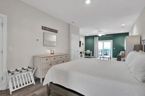A bedroom with a large bed, a dresser, a desk, and a chair. There is a sign on the wall and a view of the outside.
