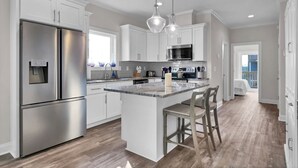 A modern kitchen with stainless steel appliances, a central island with bar stools, and a view to a balcony. - Coastal Retreat: Elevator, Pool now open, Walk to Beach! (North Topsail Beach)