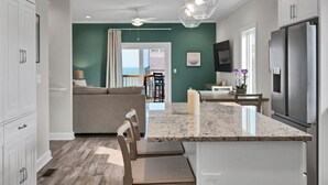 A modern kitchen with a large island, a dining area with a view, and a living room with a sofa and a TV. - Coastal Retreat: Elevator, Pool now open, Walk to Beach! (North Topsail Beach)