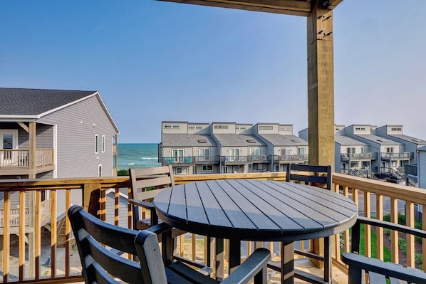 A wooden deck with a round table and chairs, overlooking a beachfront with modern buildings.