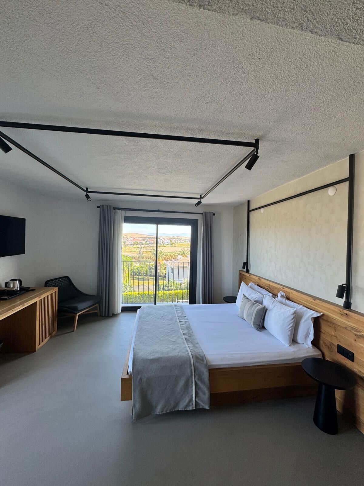 A modern hotel room with a large bed, a desk, a chair, a TV, and a balcony with a city view.