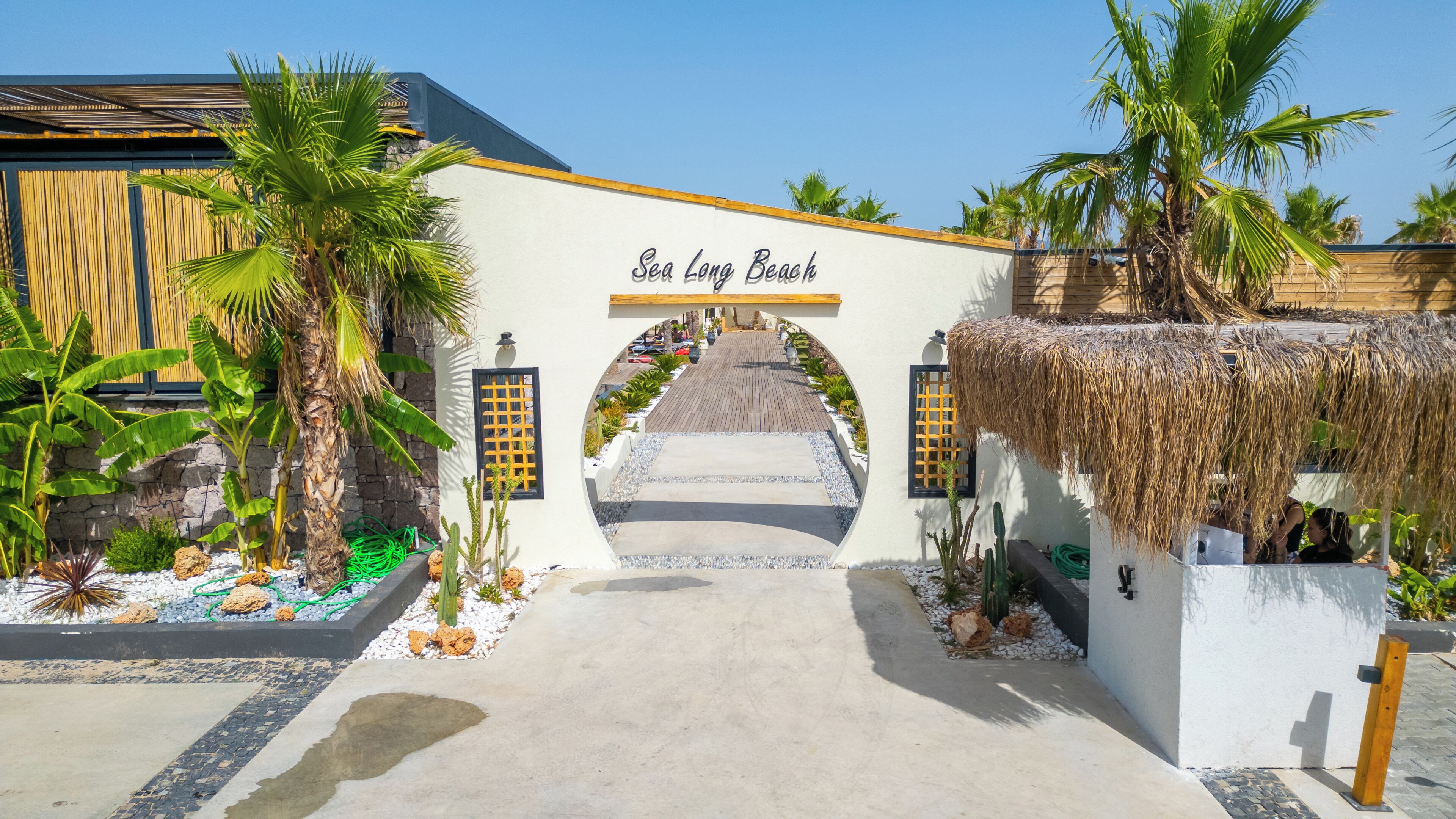 A tropical resort entrance with a sign reading 'Sea Long Beach,' palm trees, a wooden pathway, and a thatched roof structure.