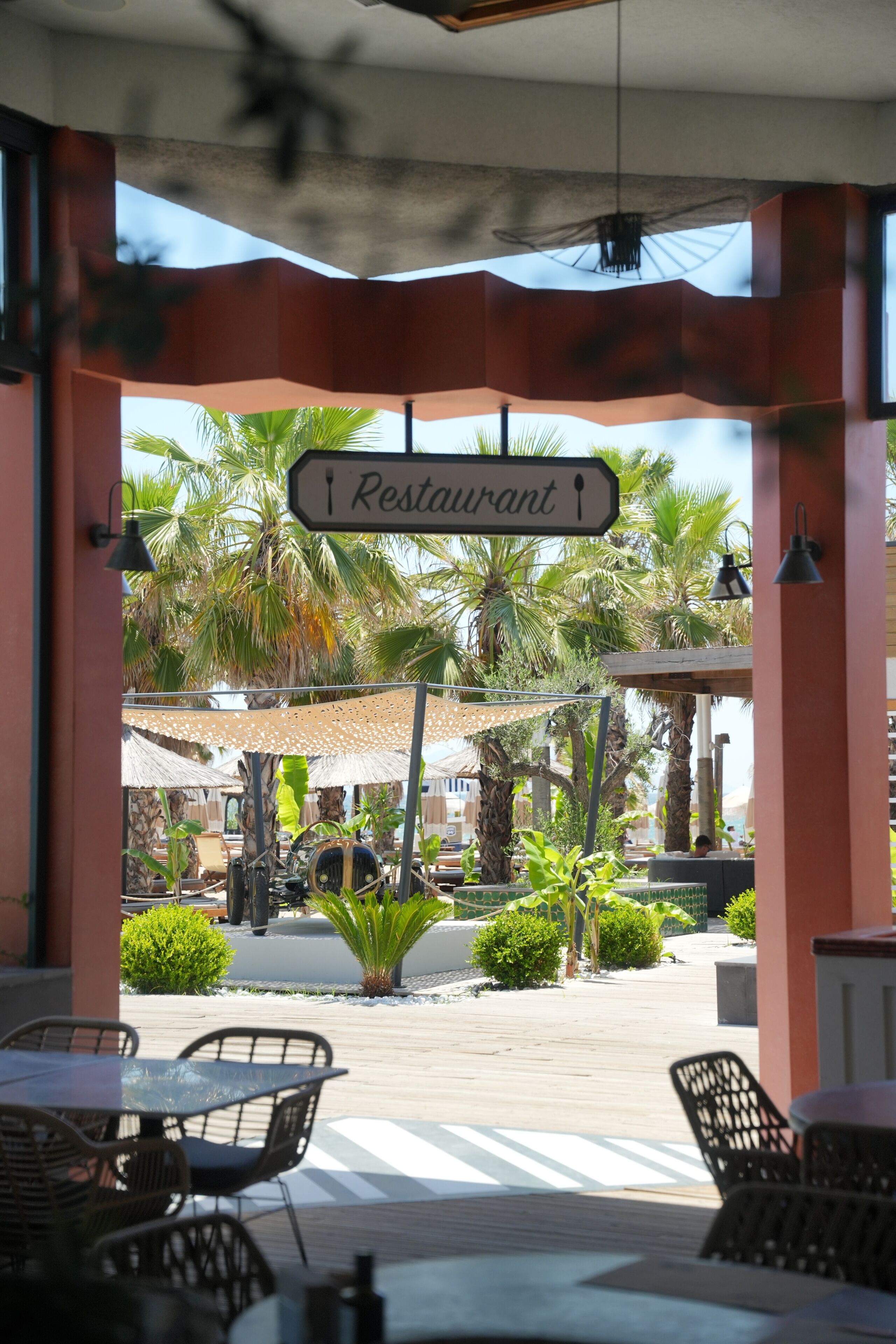 A restaurant entrance with a sign, outdoor seating, palm trees, and a covered area.