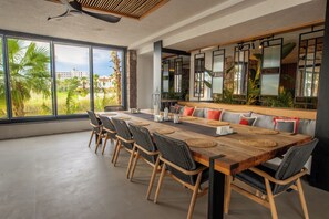 Un comedor amplio con una larga mesa de madera, sillas tejidas negras y un banco con cojines. Amplias ventanas ofrecen vistas a zonas verdes y edificios.