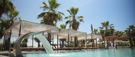 A pool with a water feature, surrounded by palm trees and covered seating areas.