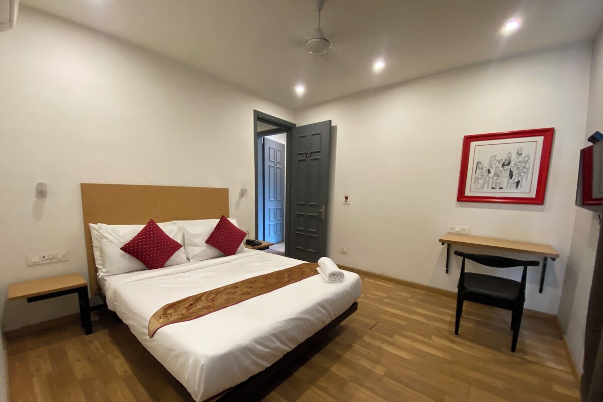 A bedroom with a bed, wooden floor, a desk, a chair, a framed picture, and a door.
