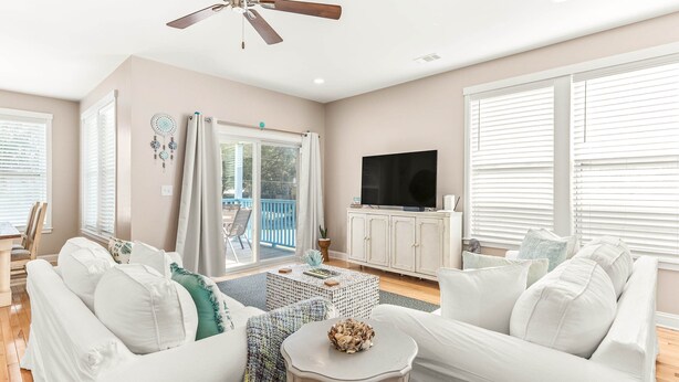 A living room with white furniture, a ceiling fan, a TV, and a sliding glass door leading to a balcony. - Peach Coral | Mid Island, Game Room, Walk to Beach (Tybee Island)