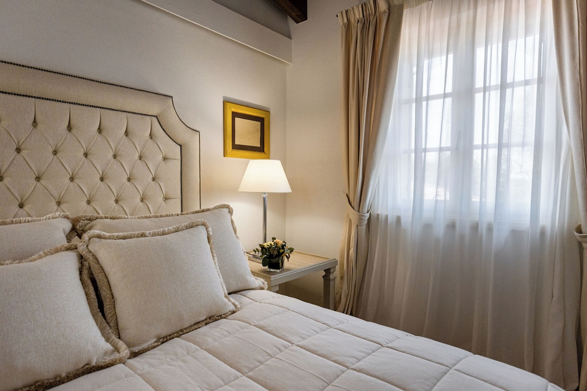 A neatly made bed with a tufted headboard, a bedside table with a lamp, and a window with sheer curtains.