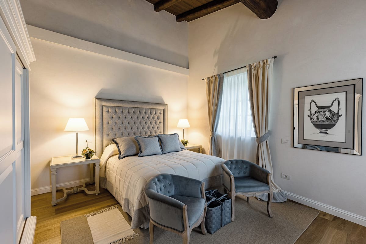A bedroom with a large bed, two bedside tables with lamps, a headboard, two armchairs, a rug, and a framed picture on the wall.