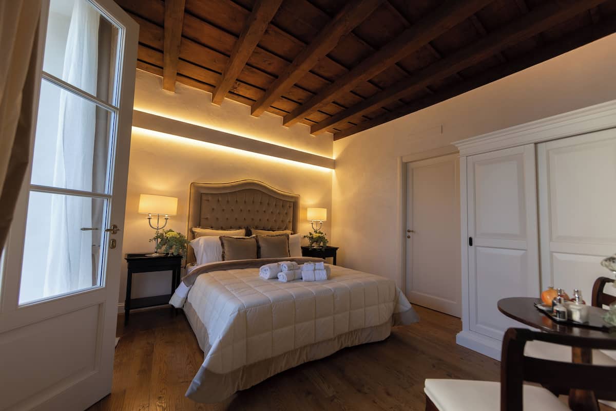 A bedroom with a large bed, wooden ceiling beams, a bedside table with a lamp, and a door.