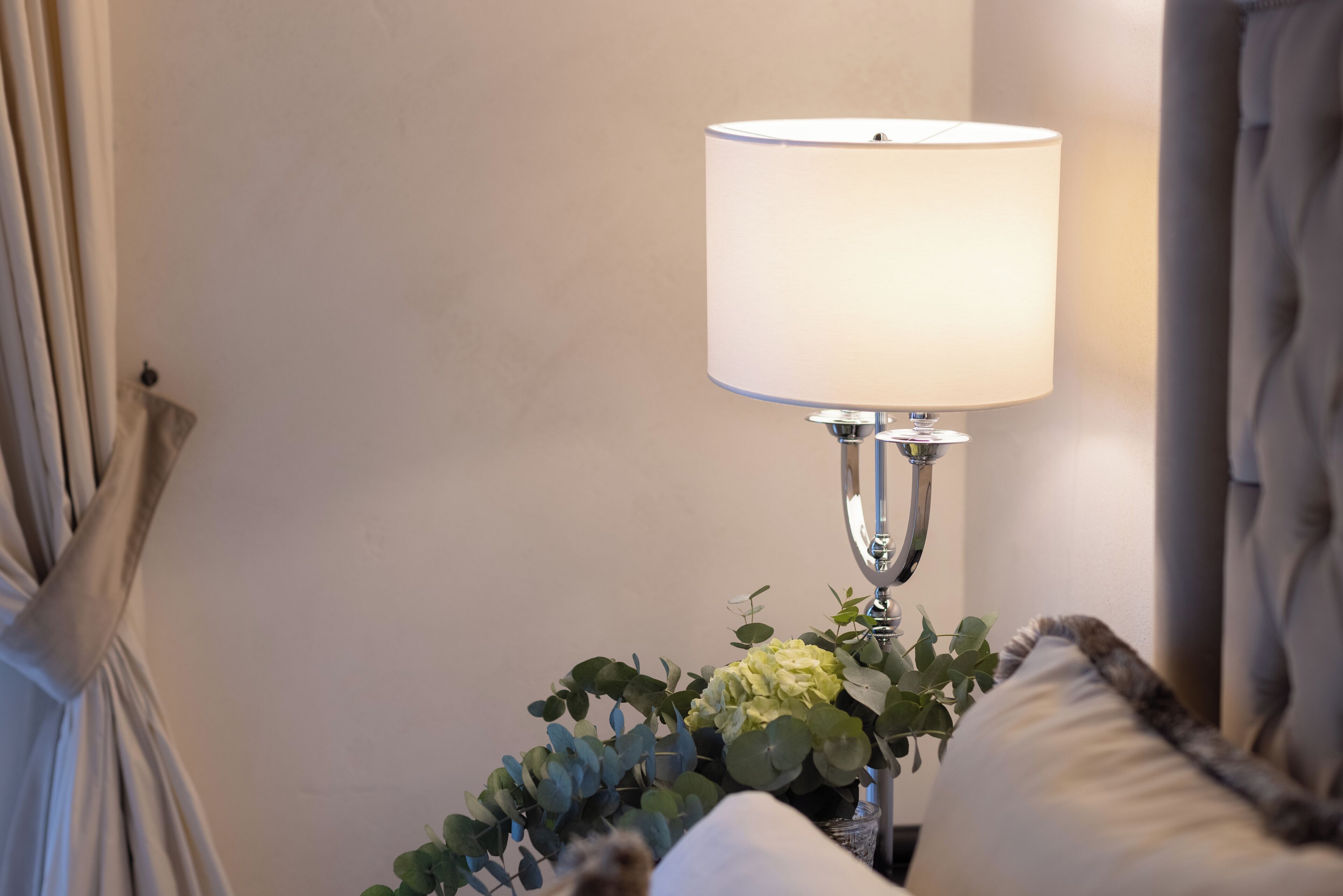 A lamp with a white shade, a cushion, and a plant on a bedside table.