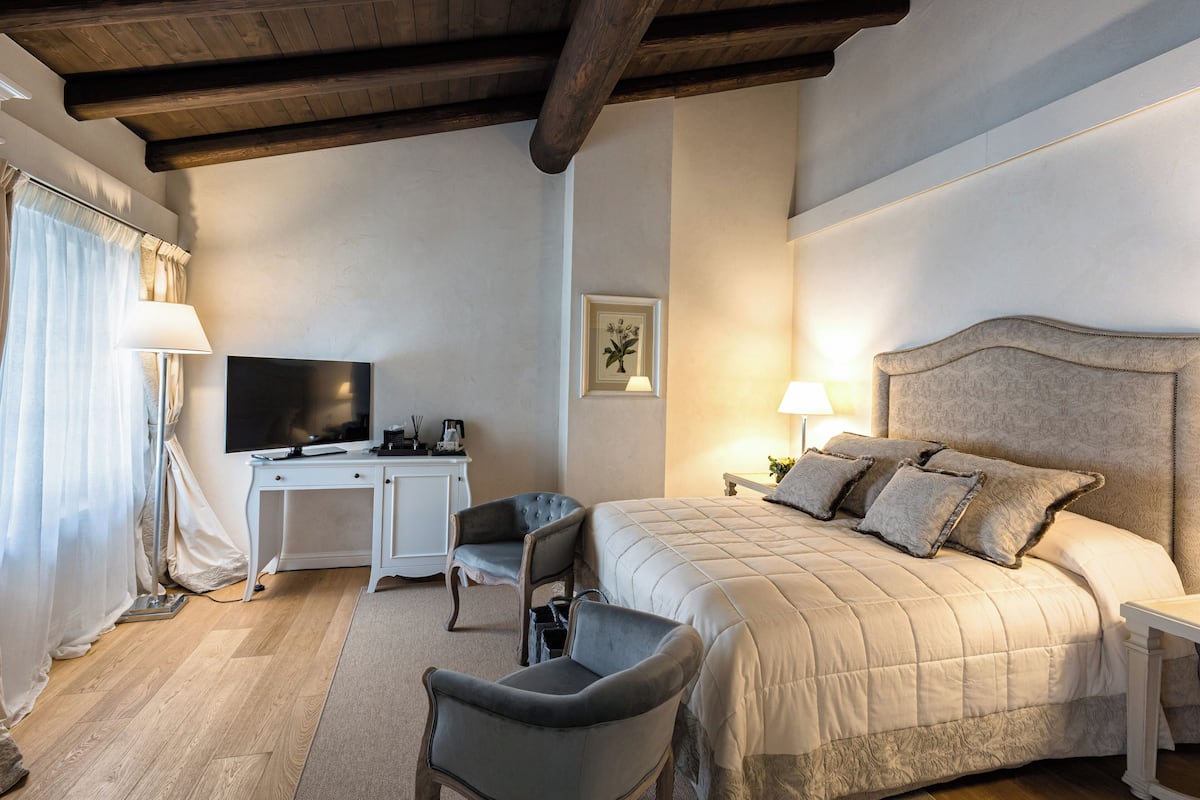 A bedroom with a large bed, a desk with a flat-screen TV, two armchairs, and a ceiling with exposed beams.