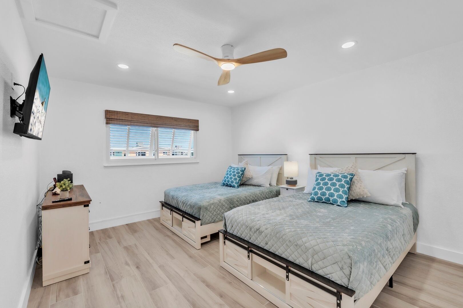 A bedroom with two beds, a ceiling fan, a window with blinds, a desk with a lamp, and a TV mounted on the wall.