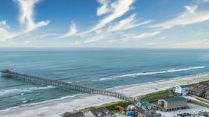 Een strand met een pier die zich uitstrekt over de oceaan, zandstrand en enkele gebouwen op de voorgrond.
