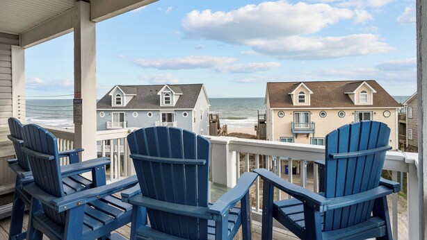 Een balkon met drie blauwe Adirondack-stoelen, met uitzicht op een strand en huizen op de achtergrond.
