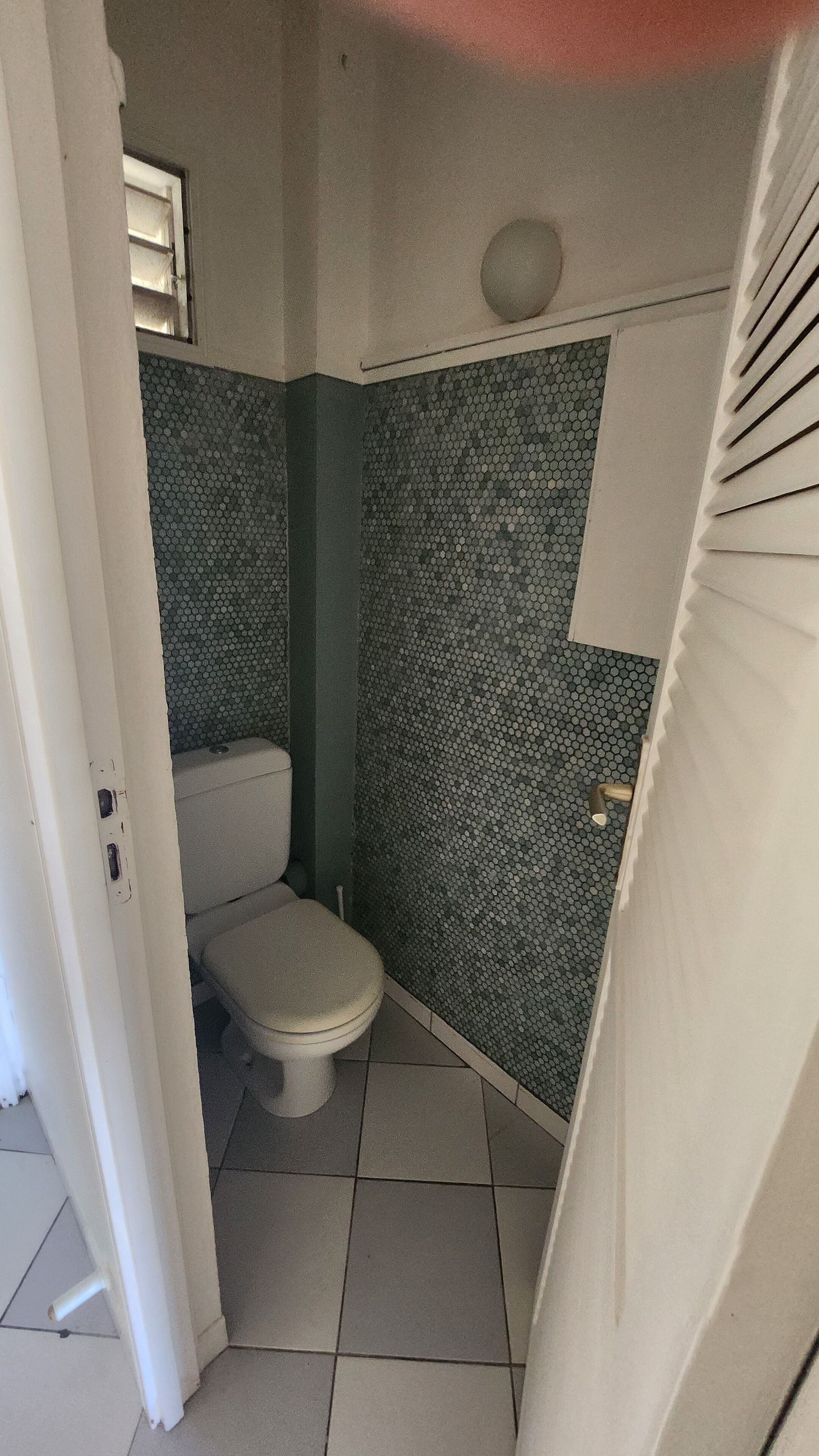 A small, narrow bathroom with a toilet and a shower area. The walls are tiled with a hexagonal pattern.