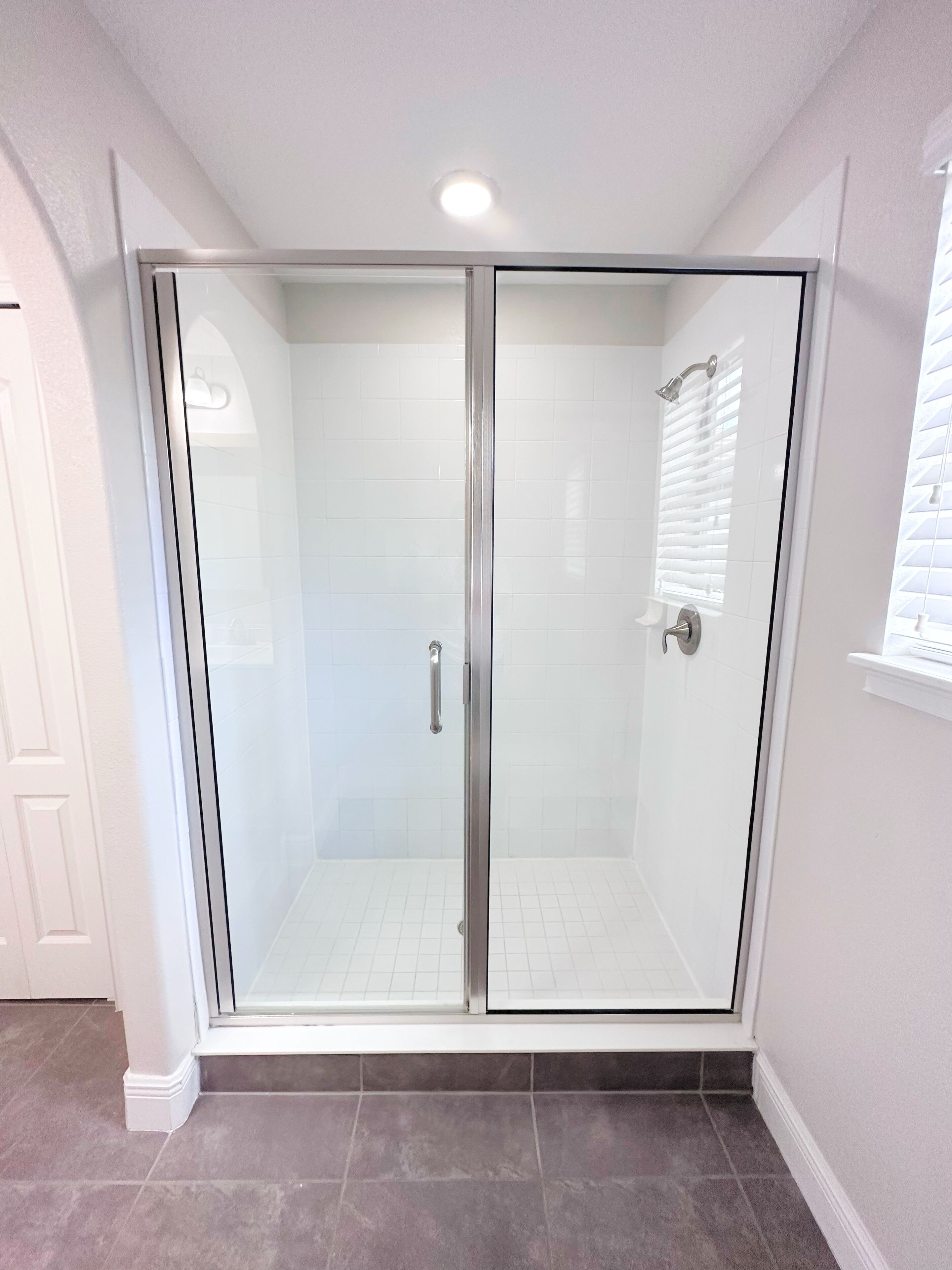 A modern shower enclosure with a glass door, a showerhead, and a window with blinds.