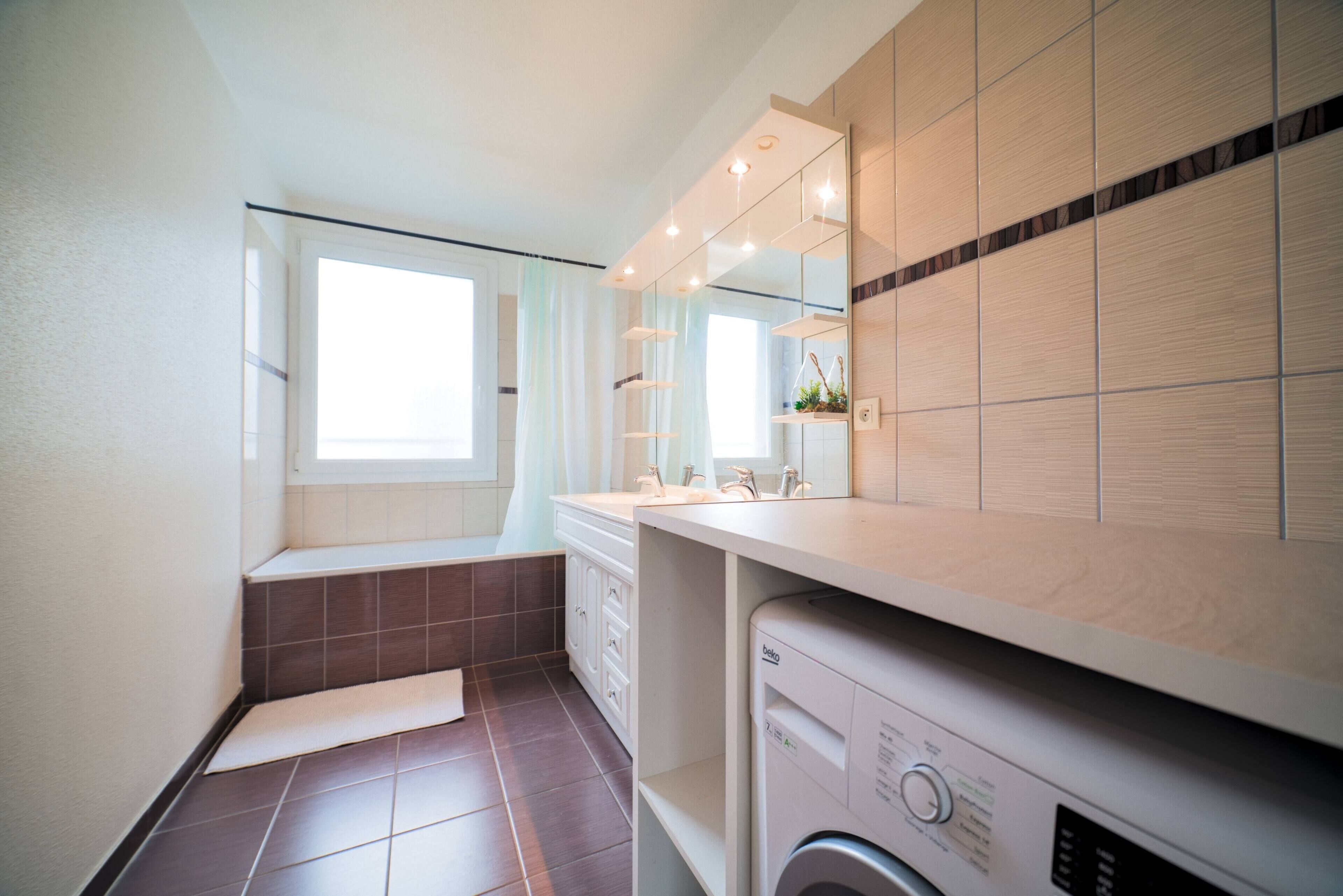 Une salle de bain moderne équipée d’un lave-linge intégré, d’un grand miroir et d’une fenêtre avec des rideaux.