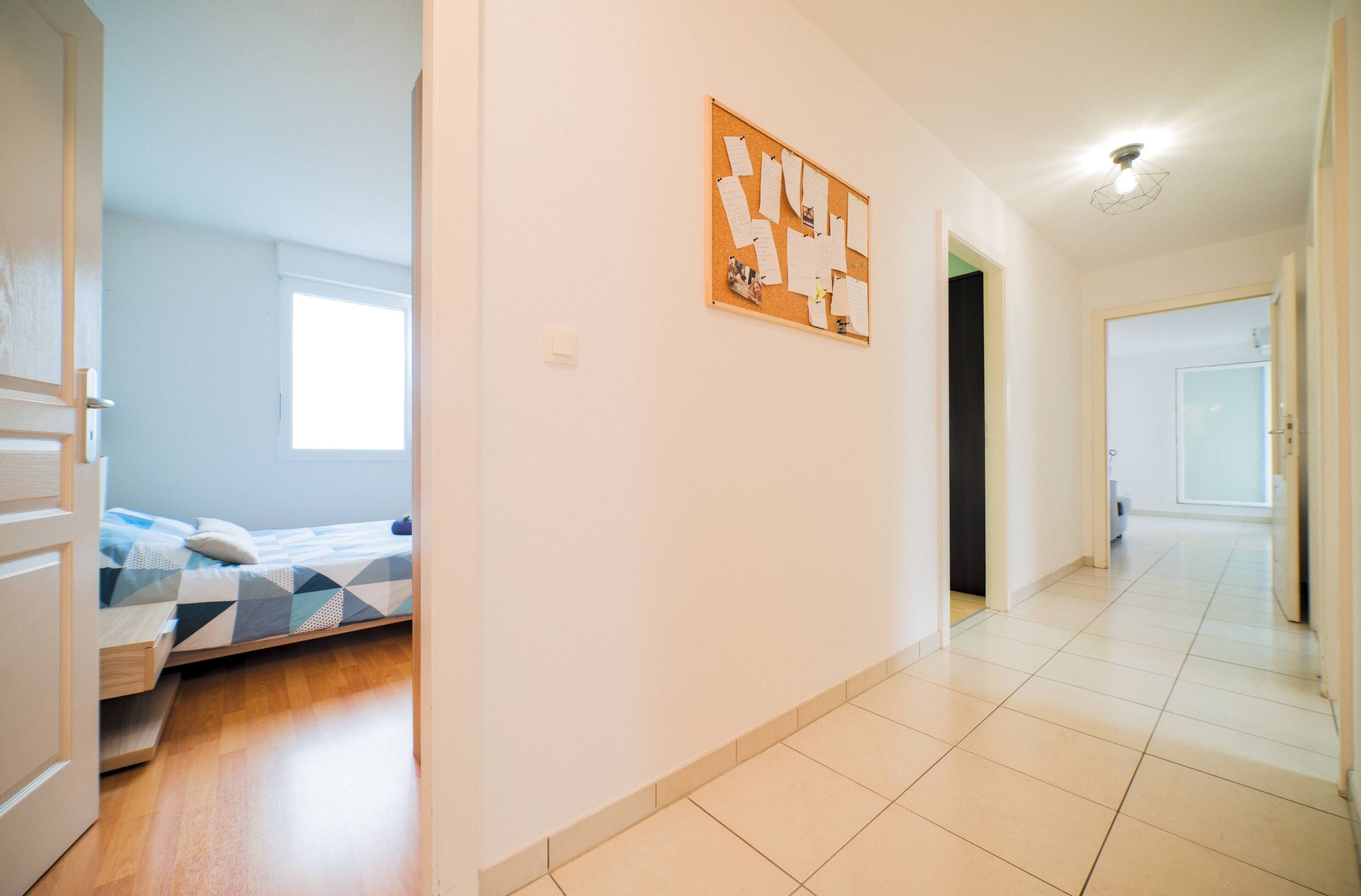 A modern, well-lit room with a bed, a corkboard with notes, and a hallway leading to another room.