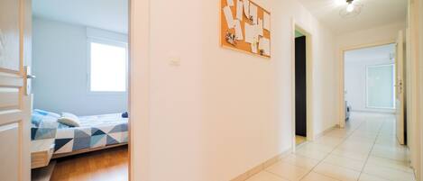 A modern, well-lit room with a bed, a corkboard with notes, and a hallway leading to another room.