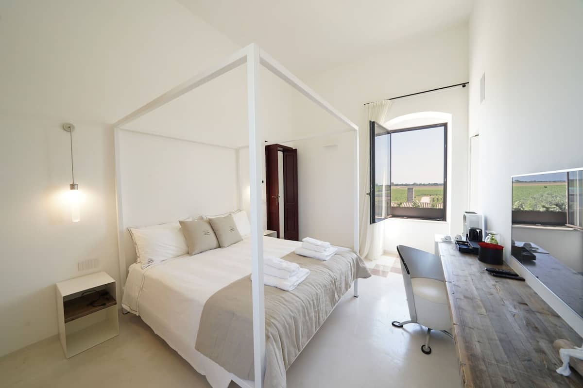 A modern bedroom with a four-poster bed, a desk with a chair, and a view of the countryside.