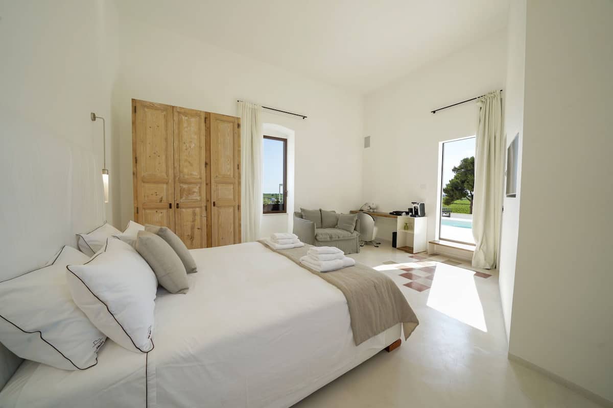 A spacious bedroom with a large bed, a wooden wardrobe, a seating area with a sofa and armchairs, and a view of the outdoors.