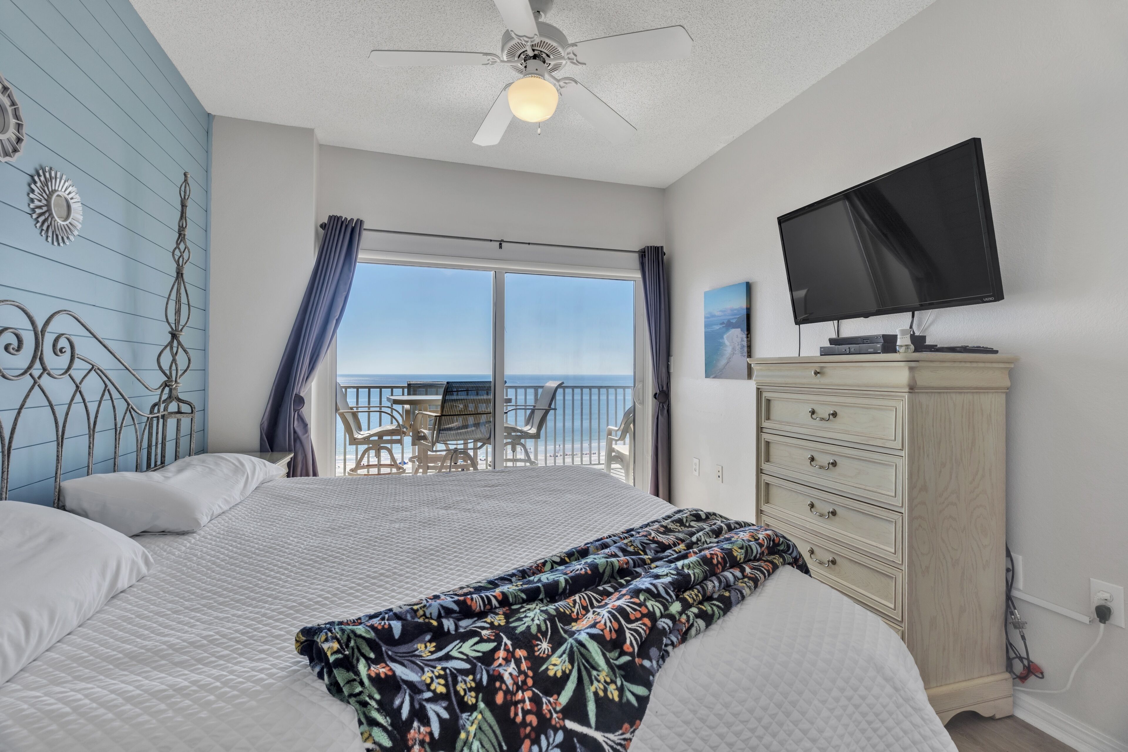 A bedroom with a bed, a television, a dresser, and a balcony with a view of the ocean.