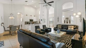 A spacious living area with a black leather sofa, a coffee table, and a dining table with chairs. There is a kitchen area with white cabinets and a microwave. The room has a high ceiling with a ceiling fan and pendant lights. - Last Minute Disney! Private Pool, Game Room & Themed Bedrooms. Book Now! #93821 (Orlando)