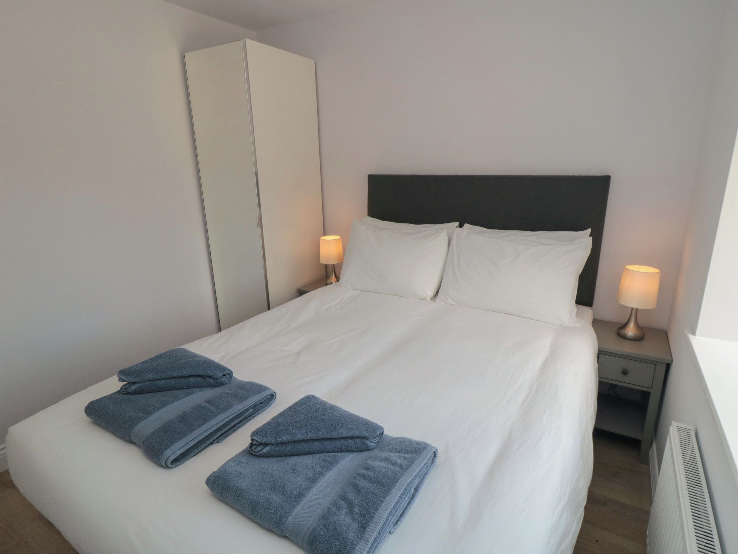 Un lit bien fait, avec du linge de lit blanc et deux serviettes pliées, une table de chevet avec une lampe et une armoire dans une chambre d’hôtel.