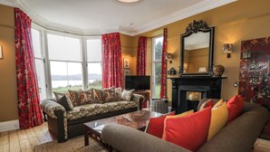 Ein Wohnzimmer mit Sofa, Couchtisch und Kamin. Ein großes Fenster mit Vorhängen, ein Spiegel und eine Lampe sind ebenfalls vorhanden.