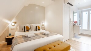 A modern bedroom with a large bed, a bench, a bedside table, and a bathroom visible through an open door.