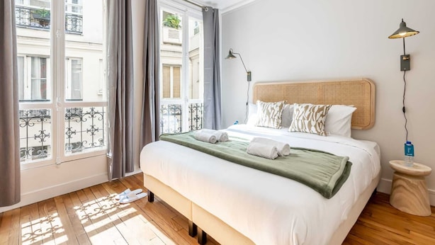 Een slaapkamer met een groot bed, een nachtkastje, een lamp en uitzicht naar buiten door een raam met gordijnen.