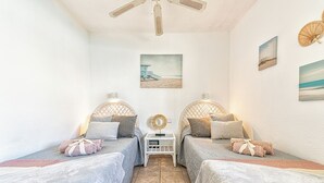 A bedroom with two beds, each with a headboard, a bedside table, and a wall-mounted lamp. There is a ceiling fan, a painting on the wall, and a beach scene picture.