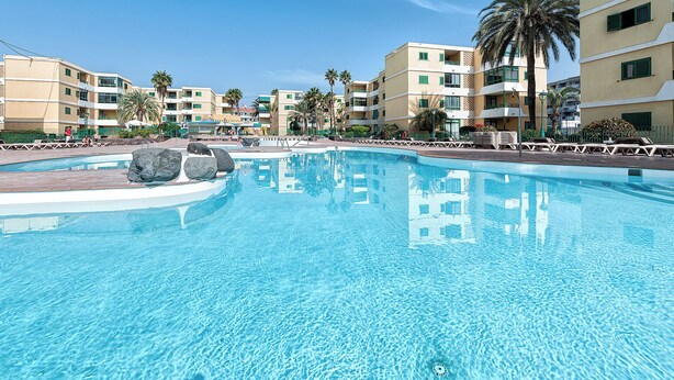 A large swimming pool with clear blue water, surrounded by multi-story buildings with balconies and palm trees.