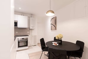 A modern kitchen with white cabinets, a built-in oven, and a dining area with a black table and chairs. - Sweett | Wagram Deluxe III (Paris)