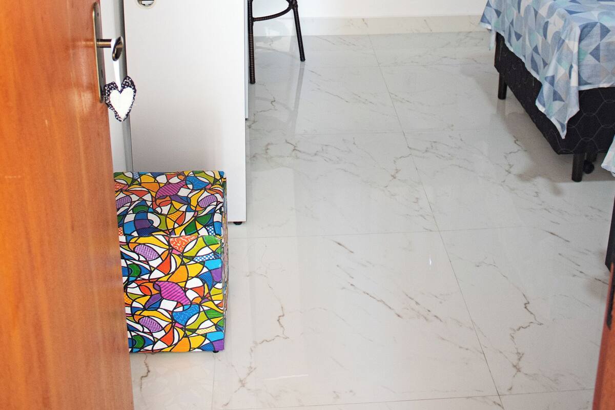 Une pièce de taille modeste comprenant un lit, un bureau avec une chaise, un petit réfrigérateur et un tapis coloré.