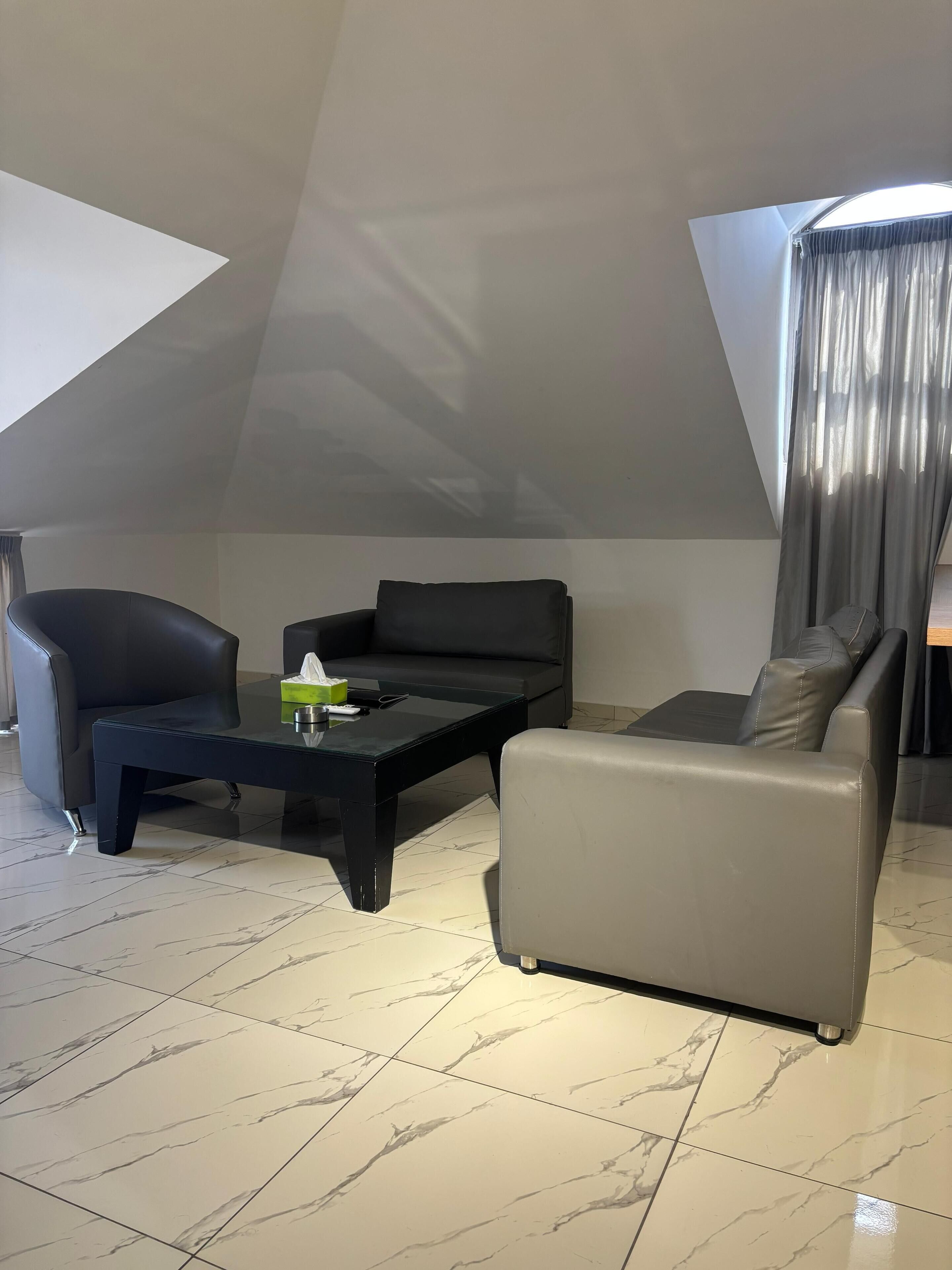 A modern loft-style living room with a high ceiling, a black leather sofa, a grey sofa, and a glass coffee table.