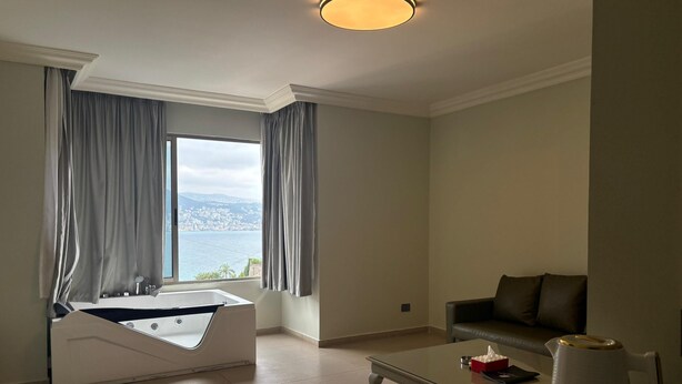 A modern bathroom with a bathtub, a large window with curtains, and a view of a cityscape. - Hotel San Diego (Kfar Yassine)