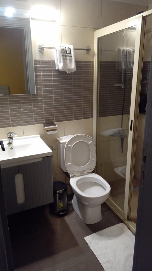 Un baño con lavamanos, inodoro y ducha blancos. Hay toallas colgadas en un perchero y un cubo de basura.
